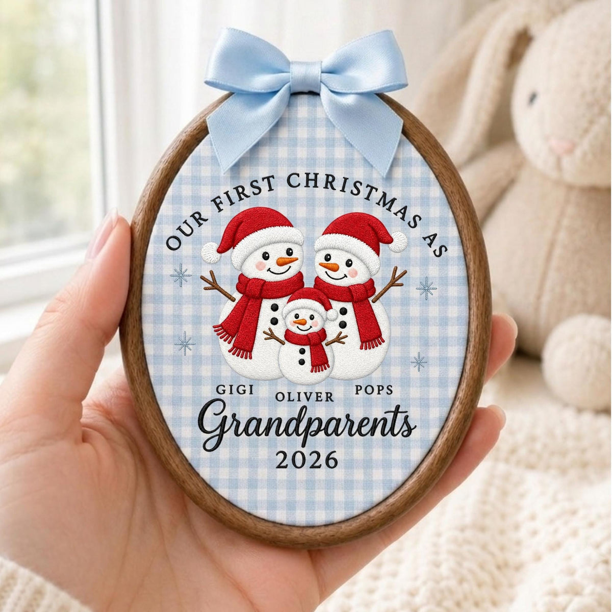May include: An oval wooden embroidery hoop ornament featuring a light blue gingham fabric background. It displays three embroidered snowmen wearing red hats and scarves, with the text Our First Christmas as Gigi, Oliver, Pops Grandparents 2026.