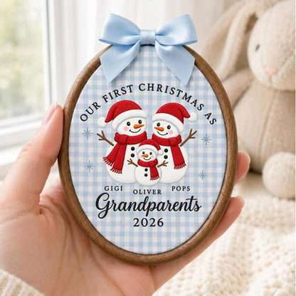 May include: An oval wooden embroidery hoop ornament featuring a light blue gingham fabric background. It displays three embroidered snowmen wearing red hats and scarves, with the text Our First Christmas as Gigi, Oliver, Pops Grandparents 2026.