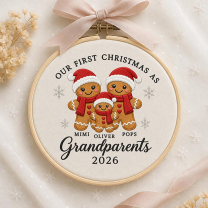 May include: A round wooden embroidery hoop containing white fabric with a stitched design of three gingerbread figures wearing red hats and scarves. The text reads: OUR FIRST CHRISTMAS AS MIMI OLIVER POPS Grandparents 2026. A light pink ribbon is tied at the top.