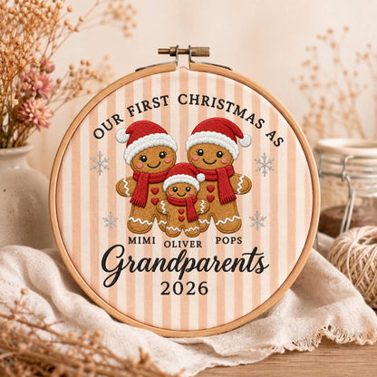 May include: A round wooden embroidery hoop containing fabric with a beige and white vertical striped pattern. The design features three embroidered gingerbread figures wearing red hats and scarves, with the text "OUR FIRST CHRISTMAS AS MIMI OLIVER POPS Grandparents 2026" and small snowflake icons.