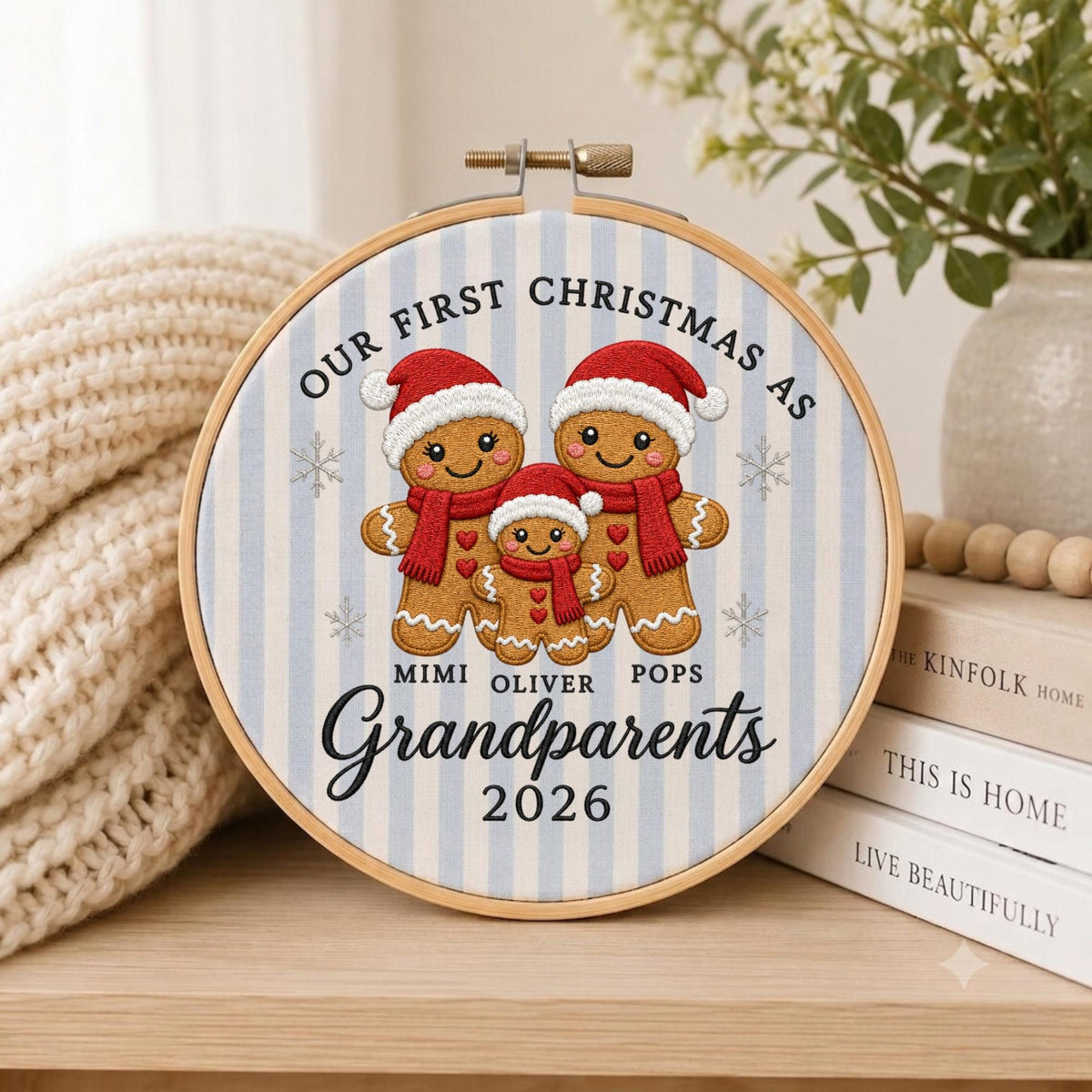 May include: A circular embroidery hoop featuring a light blue and white striped fabric background. The design displays three gingerbread figures wearing red scarves and Santa hats, with the text "OUR FIRST CHRISTMAS AS MIMI OLIVER POPS Grandparents 2026".