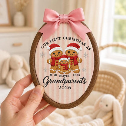 May include: An oval wooden embroidery hoop frame containing fabric with a pink and white striped pattern. The fabric features three embroidered gingerbread figures wearing Santa hats and red scarves, with the text "OUR FIRST CHRISTMAS AS MIMI OLIVER POPS Grandparents 2026" and a pink satin bow at the top.