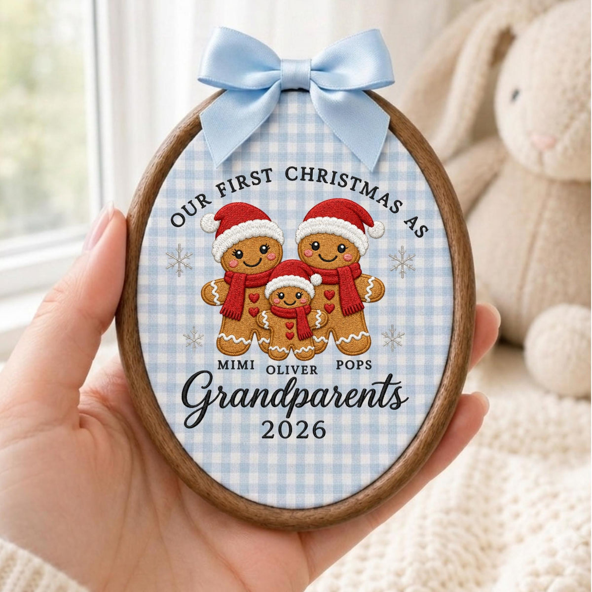May include: An oval wooden embroidery hoop ornament featuring a light blue gingham fabric background. It displays three embroidered gingerbread figures wearing red hats and scarves, with the text "OUR FIRST CHRISTMAS AS MIMI OLIVER POPS Grandparents 2026" and a light blue satin bow at the top.