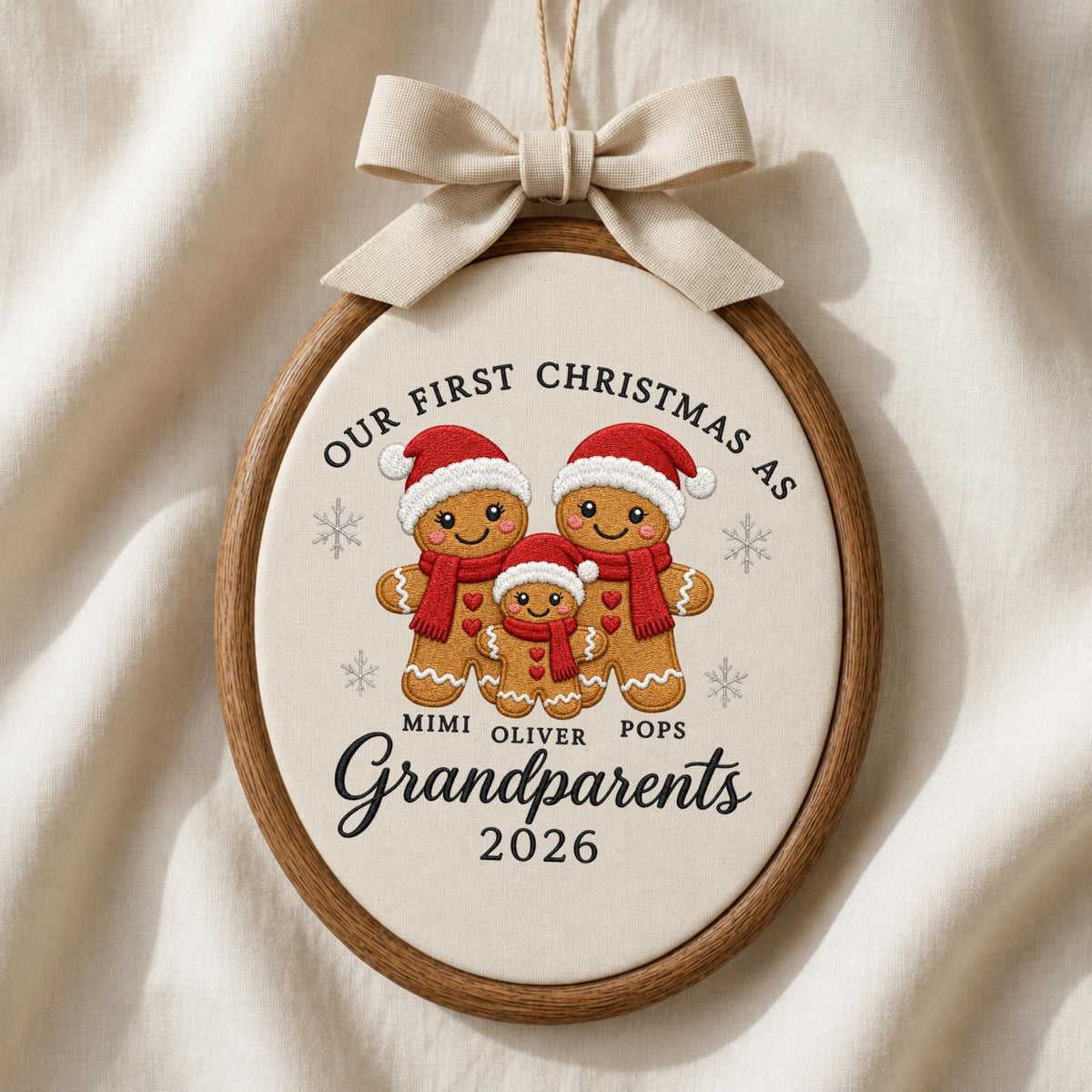 May include: An oval wooden embroidery hoop ornament featuring a cream fabric center with embroidered gingerbread figures wearing red hats and scarves. Text reads: OUR FIRST CHRISTMAS AS MIMI OLIVER POPS Grandparents 2026. A beige ribbon bow is attached at the top.