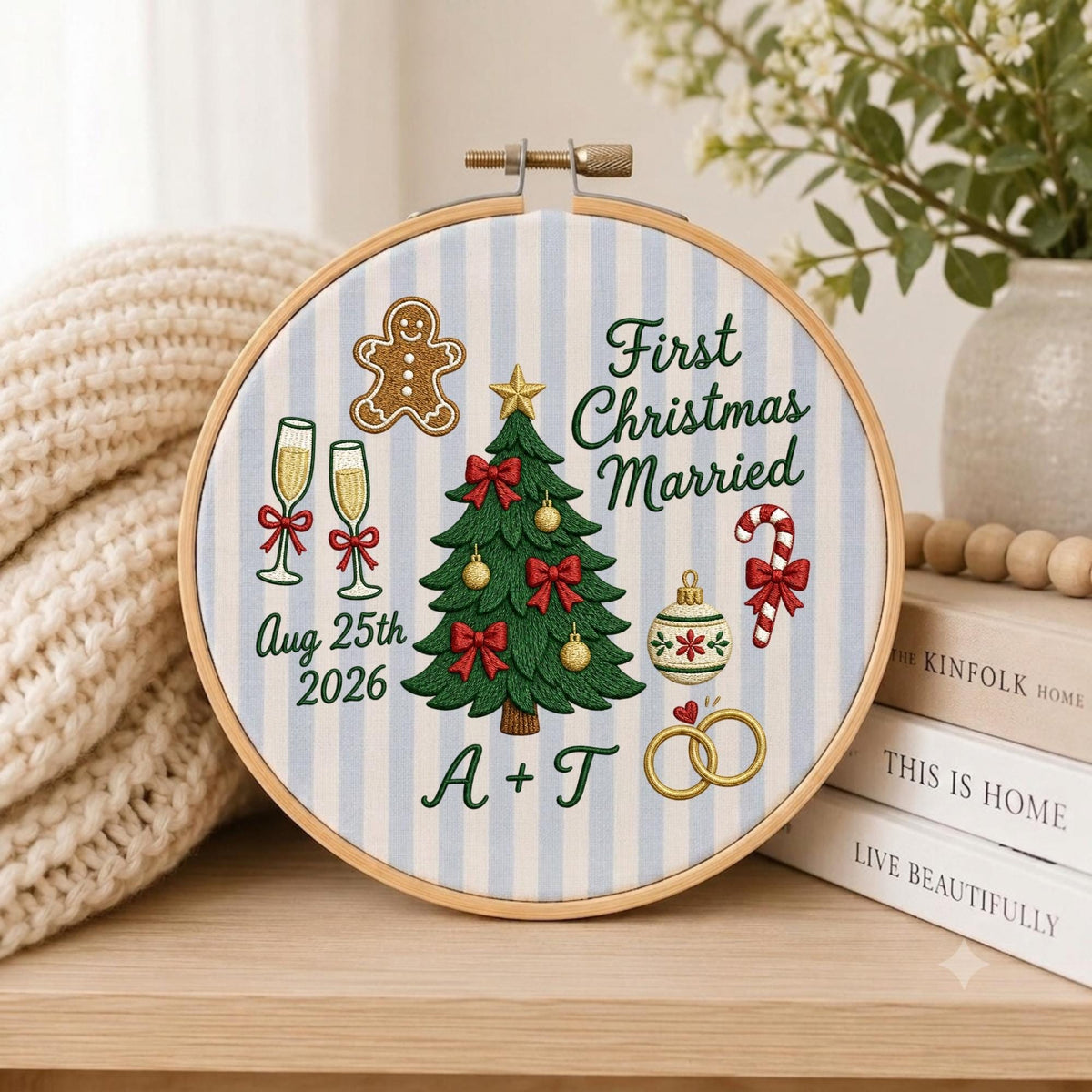 May include: A round embroidery hoop featuring a light blue and white striped fabric with a central green Christmas tree, a gingerbread man, champagne flutes, a candy cane, and two gold rings. Text reads First Christmas Married, Aug 25th 2026, and A + T.