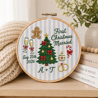 May include: A round embroidery hoop featuring a light blue and white striped fabric with a central green Christmas tree, a gingerbread man, champagne flutes, a candy cane, and two gold rings. Text reads First Christmas Married, Aug 25th 2026, and A + T.