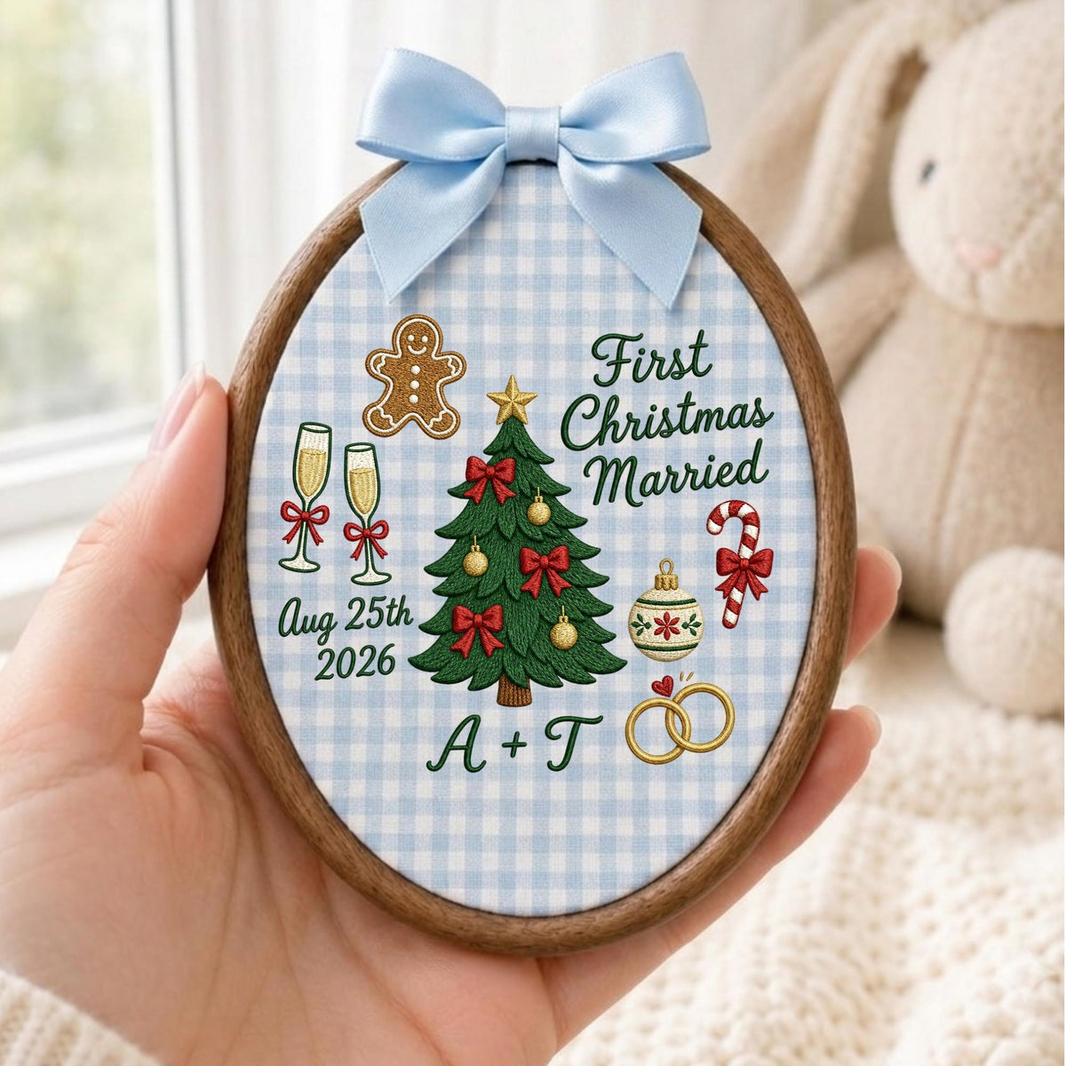 May include: An oval embroidery hoop ornament featuring a light blue and white gingham fabric background. It displays a green Christmas tree, a gingerbread man, champagne flutes, a candy cane, and gold rings. Text reads First Christmas Married, Aug 25th 2026, and A + T, topped with a light blue satin bow.