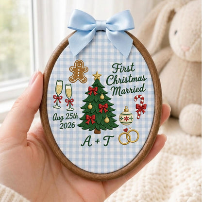 May include: An oval embroidery hoop ornament featuring a light blue and white gingham fabric background. It displays a green Christmas tree, a gingerbread man, champagne flutes, a candy cane, and gold rings. Text reads First Christmas Married, Aug 25th 2026, and A + T, topped with a light blue satin bow.