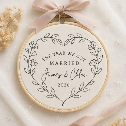 May include: A circular wooden embroidery hoop containing off-white fabric with black embroidered text that reads THE YEAR WE GOT MARRIED James &amp; Chloe 2026, framed by a black floral heart outline and topped with a light pink satin ribbon bow.