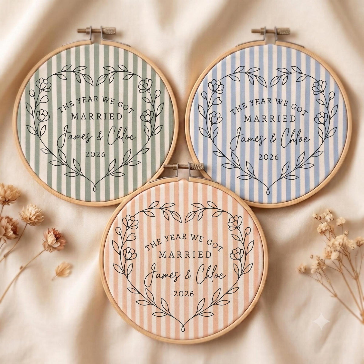 May include: Three circular wooden embroidery hoops containing fabric with vertical stripes in sage green, light blue, and peach. Each features a black floral heart outline, the text THE YEAR WE GOT MARRIED, the names James &amp; Chloe, and the year 2026.