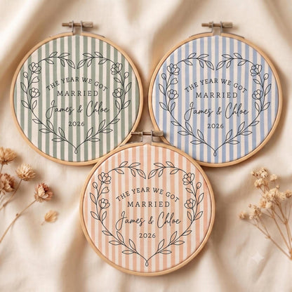 May include: Three circular wooden embroidery hoops containing fabric with vertical stripes in sage green, light blue, and peach. Each features a black floral heart outline, the text THE YEAR WE GOT MARRIED, the names James &amp; Chloe, and the year 2026.