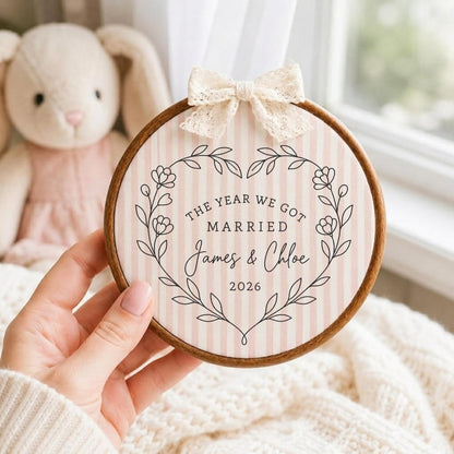 May include: A circular wooden embroidery hoop containing fabric with a light pink and white vertical striped pattern. The fabric features a black line art floral heart wreath surrounding the text: THE YEAR WE GOT MARRIED James &amp; Chloe 2026.
