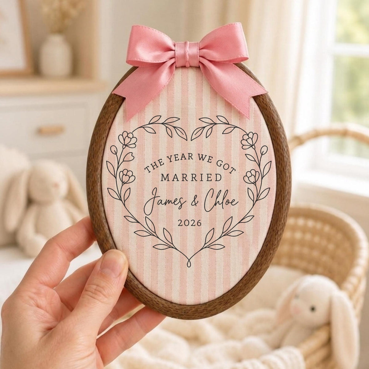 May include: An oval wooden embroidery hoop frame containing light pink and white striped fabric. A pink satin ribbon bow is attached to the top. The fabric features a black line art floral heart wreath with the text: THE YEAR WE GOT MARRIED James &amp; Chloe 2026.