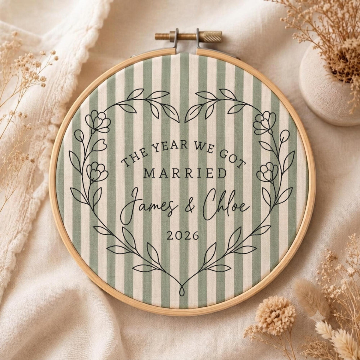 May include: A circular wooden embroidery hoop containing fabric with vertical sage green and white stripes. The fabric features black embroidered text reading THE YEAR WE GOT MARRIED James &amp; Chloe 2026, framed by a delicate floral vine wreath.