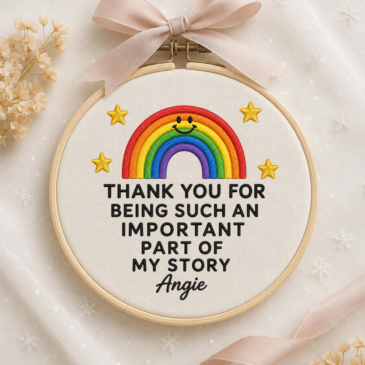 May include: A circular wooden embroidery hoop containing white fabric with a stitched rainbow featuring a smiling face, four yellow stars, and the black text: THANK YOU FOR BEING SUCH AN IMPORTANT PART OF MY STORY Angie. A light pink ribbon is tied at the top.
