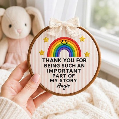 May include: A round wooden embroidery hoop containing fabric with vertical pink stripes. It features an embroidered rainbow with a smiley face, four yellow stars, and the text: THANK YOU FOR BEING SUCH AN IMPORTANT PART OF MY STORY Angie. A cream lace bow is attached to the top.