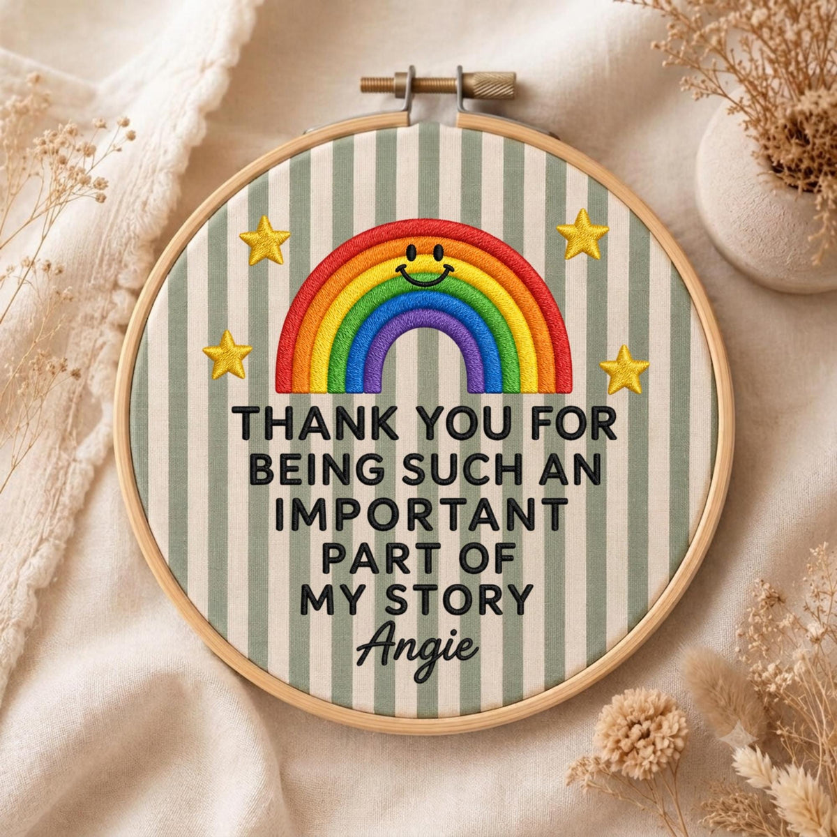 May include: A circular wooden embroidery hoop containing fabric with vertical sage green and white stripes. It features a colorful embroidered rainbow with a smiling face, four yellow stars, and the text: THANK YOU FOR BEING SUCH AN IMPORTANT PART OF MY STORY Angie.