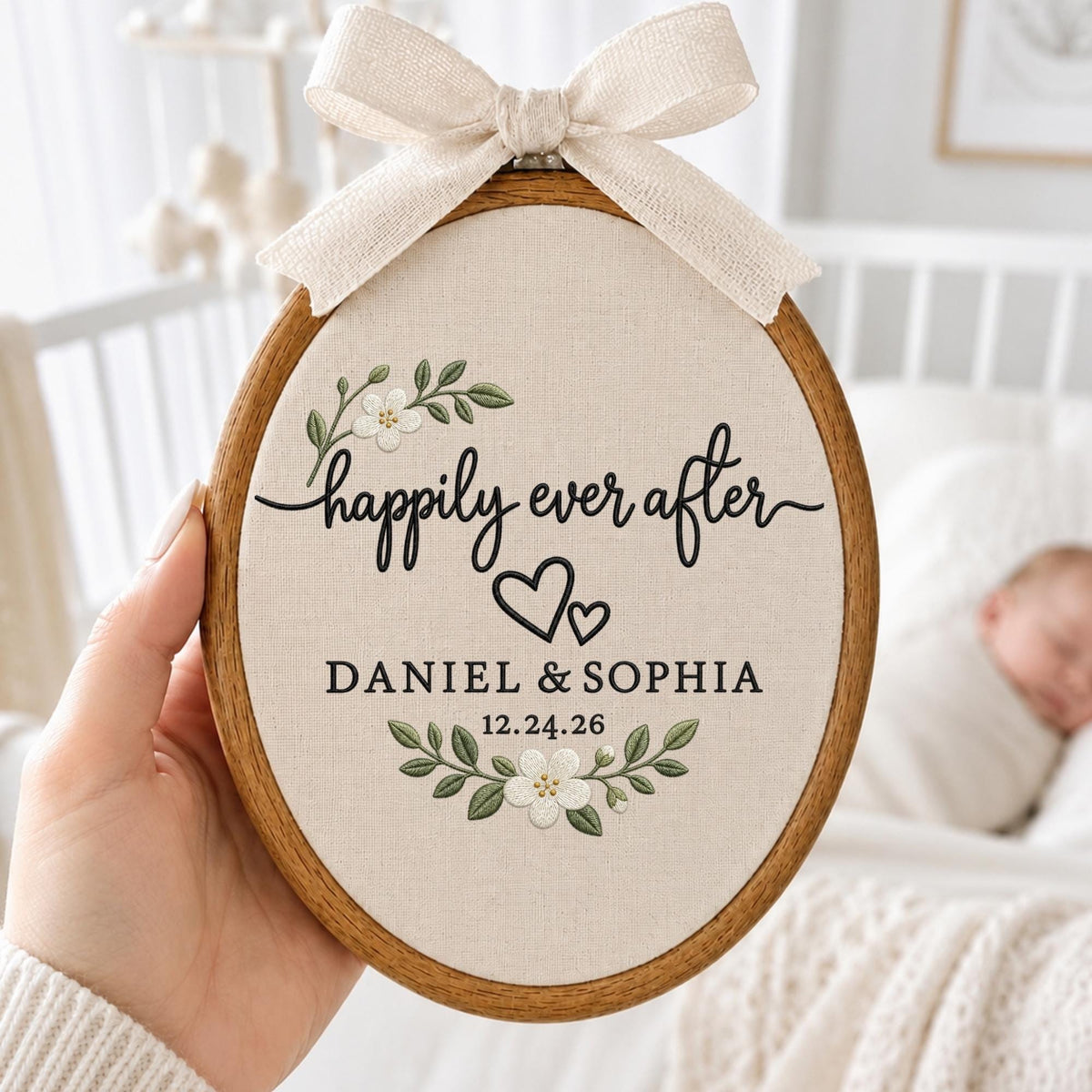 May include: An oval wooden embroidery hoop containing cream-colored fabric with the embroidered text "happily ever after", "DANIEL &amp; SOPHIA", and "12.24.26". The design features two black hearts and green floral sprigs with white flowers, topped with a beige ribbon bow.