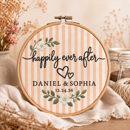 May include: A round wooden embroidery hoop containing fabric with vertical peach and white stripes. The fabric features black embroidered text reading "happily ever after", "DANIEL &amp; SOPHIA", and "12.24.26", accented by two white flowers and two hearts.