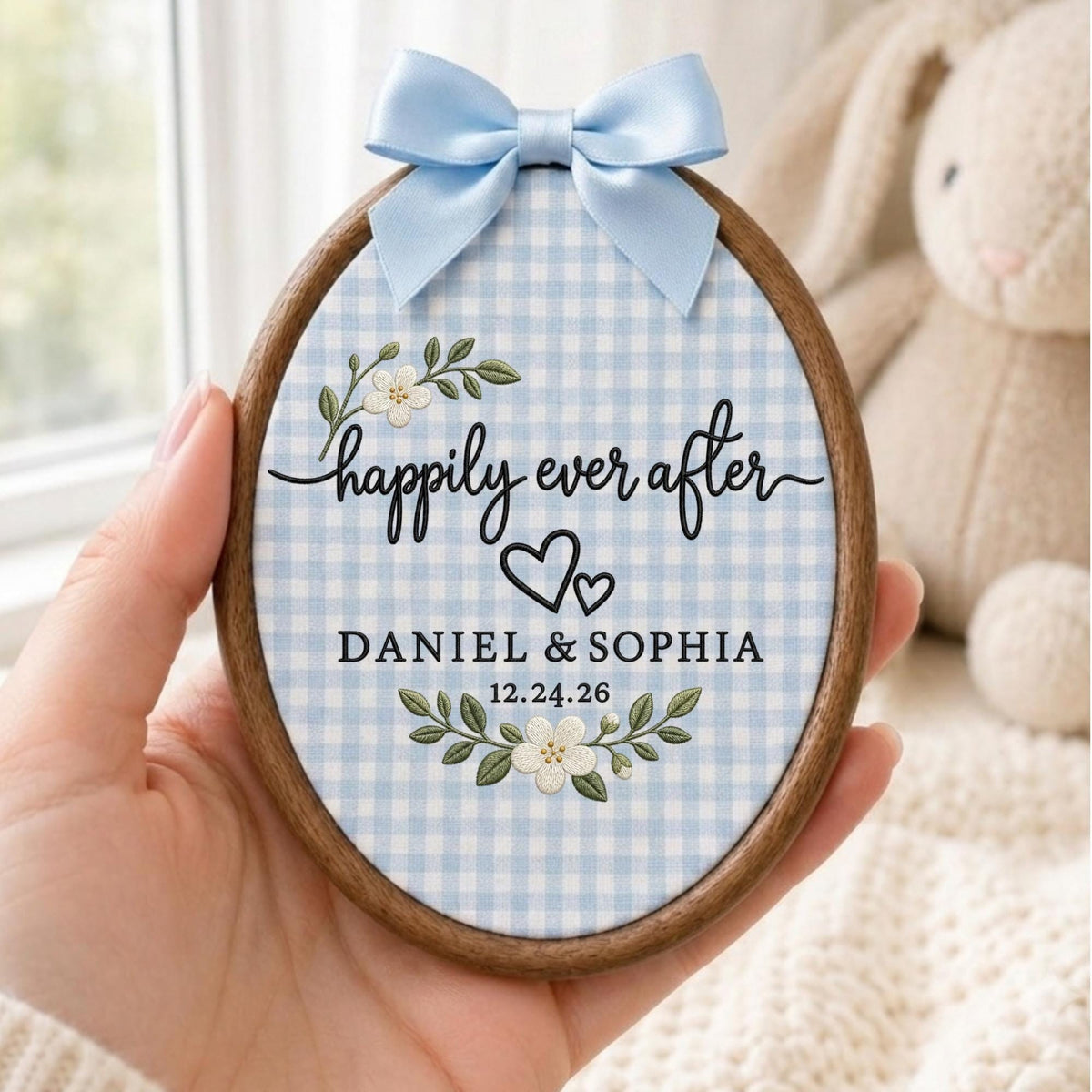May include: An oval wooden embroidery hoop frame containing light blue and white gingham fabric. The fabric features embroidered white flowers, green leaves, two black hearts, and the text: "happily ever after", "DANIEL &amp; SOPHIA", and "12.24.26". A light blue satin ribbon bow is attached to the top.