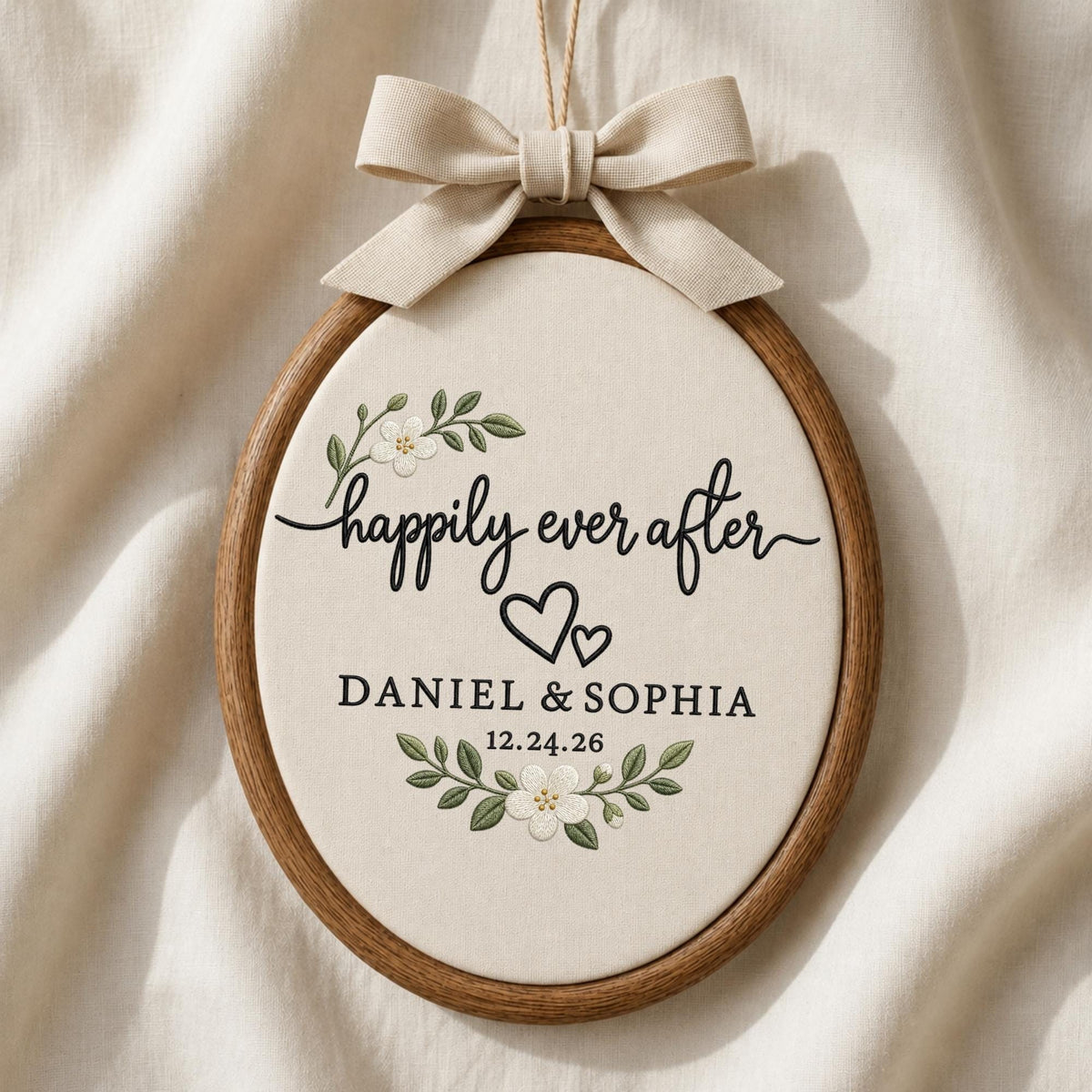 May include: An oval wooden embroidery hoop containing cream fabric with the embroidered text "happily ever after", two black hearts, the names "DANIEL &amp; SOPHIA", and the date "12.24.26", accented by floral sprigs and a beige fabric bow.