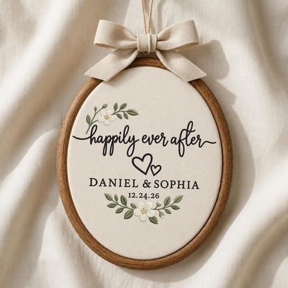 May include: An oval wooden embroidery hoop containing cream fabric with the embroidered text "happily ever after", two black hearts, the names "DANIEL &amp; SOPHIA", and the date "12.24.26", accented by floral sprigs and a beige fabric bow.