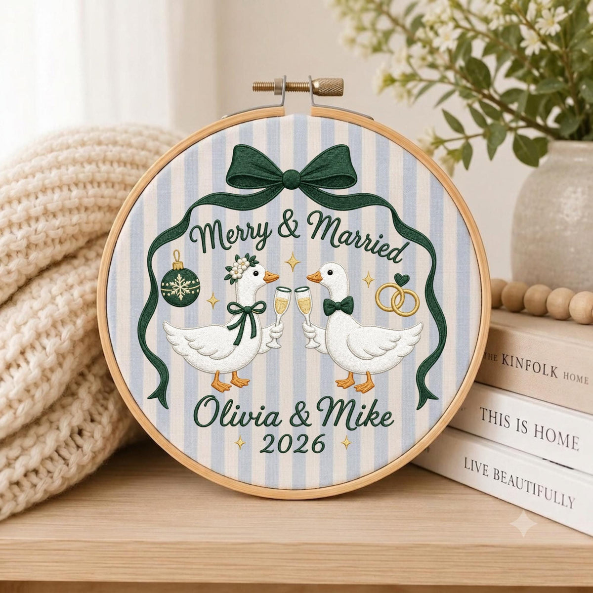 May include: A circular embroidery hoop featuring a light blue and white striped fabric background. The design displays two white geese clinking champagne glasses, a dark green bow, a Christmas ornament, two gold rings, and the text Merry &amp; Married Olivia &amp; Mike 2026.