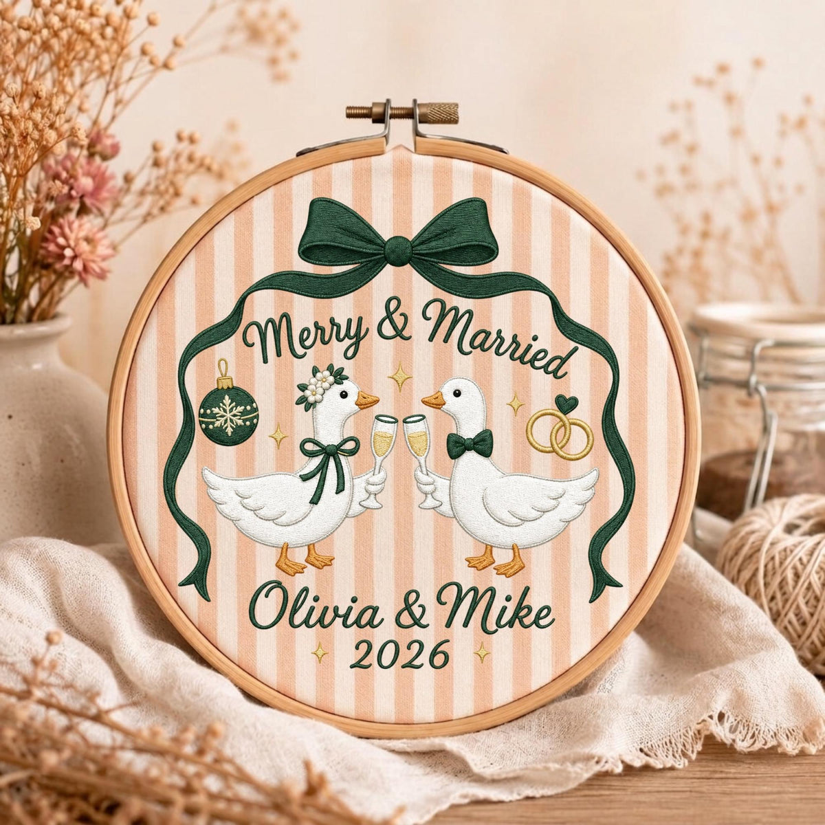 May include: A round wooden embroidery hoop containing fabric with vertical peach and white stripes. The embroidery features two white geese holding champagne flutes, a dark green bow, a Christmas ornament, wedding rings, and the text Merry &amp; Married Olivia &amp; Mike 2026.