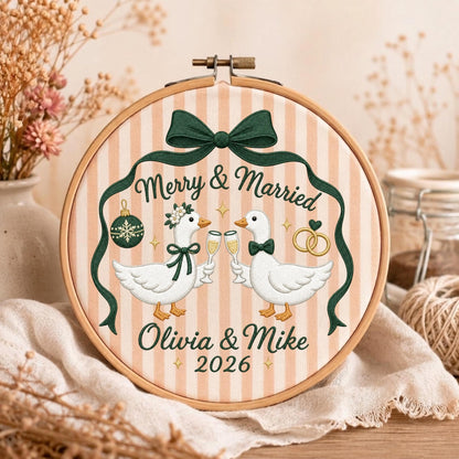 May include: A round wooden embroidery hoop containing fabric with vertical peach and white stripes. The embroidery features two white geese holding champagne flutes, a dark green bow, a Christmas ornament, wedding rings, and the text Merry &amp; Married Olivia &amp; Mike 2026.