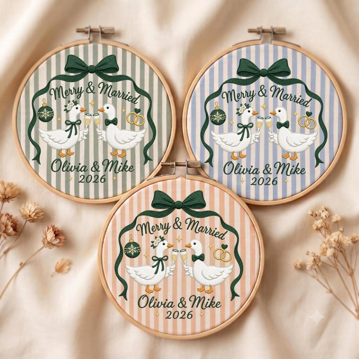 May include: Three circular embroidery hoops featuring striped fabric backgrounds in sage green, light blue, and peach. Each hoop displays two white geese clinking champagne glasses, a dark green bow, a Christmas ornament, and the text Merry &amp; Married Olivia &amp; Mike 2026.