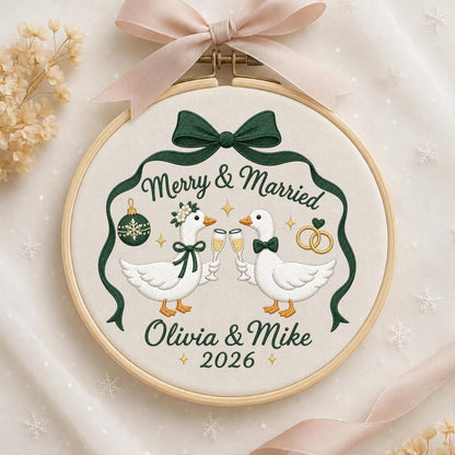 May include: An embroidery hoop featuring a cream fabric with two white geese holding champagne glasses. The text reads Merry &amp; Married, Olivia &amp; Mike, and 2026. It includes a dark green ribbon design, a Christmas ornament, and two gold rings.