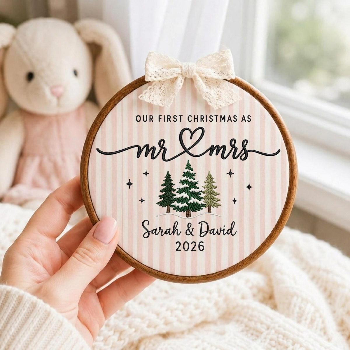 May include: A round wooden embroidery hoop frame containing fabric with a light pink and white striped pattern. The text reads OUR FIRST CHRISTMAS AS mr &amp; mrs, Sarah &amp; David, 2026. It features three embroidered pine trees and a cream lace bow.