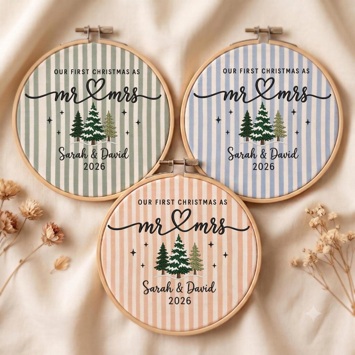 May include: Three circular embroidery hoops featuring striped fabric in sage green, light blue, and peach. Each hoop displays the text "OUR FIRST CHRISTMAS AS mr &amp; mrs", "Sarah &amp; David", and "2026" above a graphic of three snow-covered pine trees.