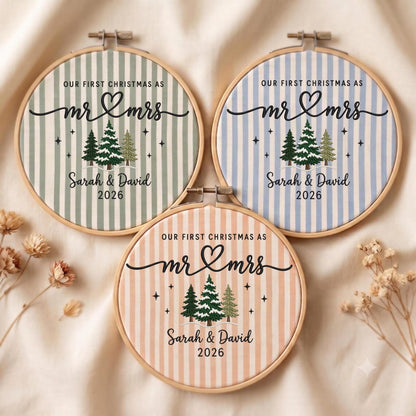 May include: Three circular embroidery hoops featuring striped fabric in sage green, light blue, and peach. Each hoop displays the text "OUR FIRST CHRISTMAS AS mr &amp; mrs", "Sarah &amp; David", and "2026" above a graphic of three snow-covered pine trees.