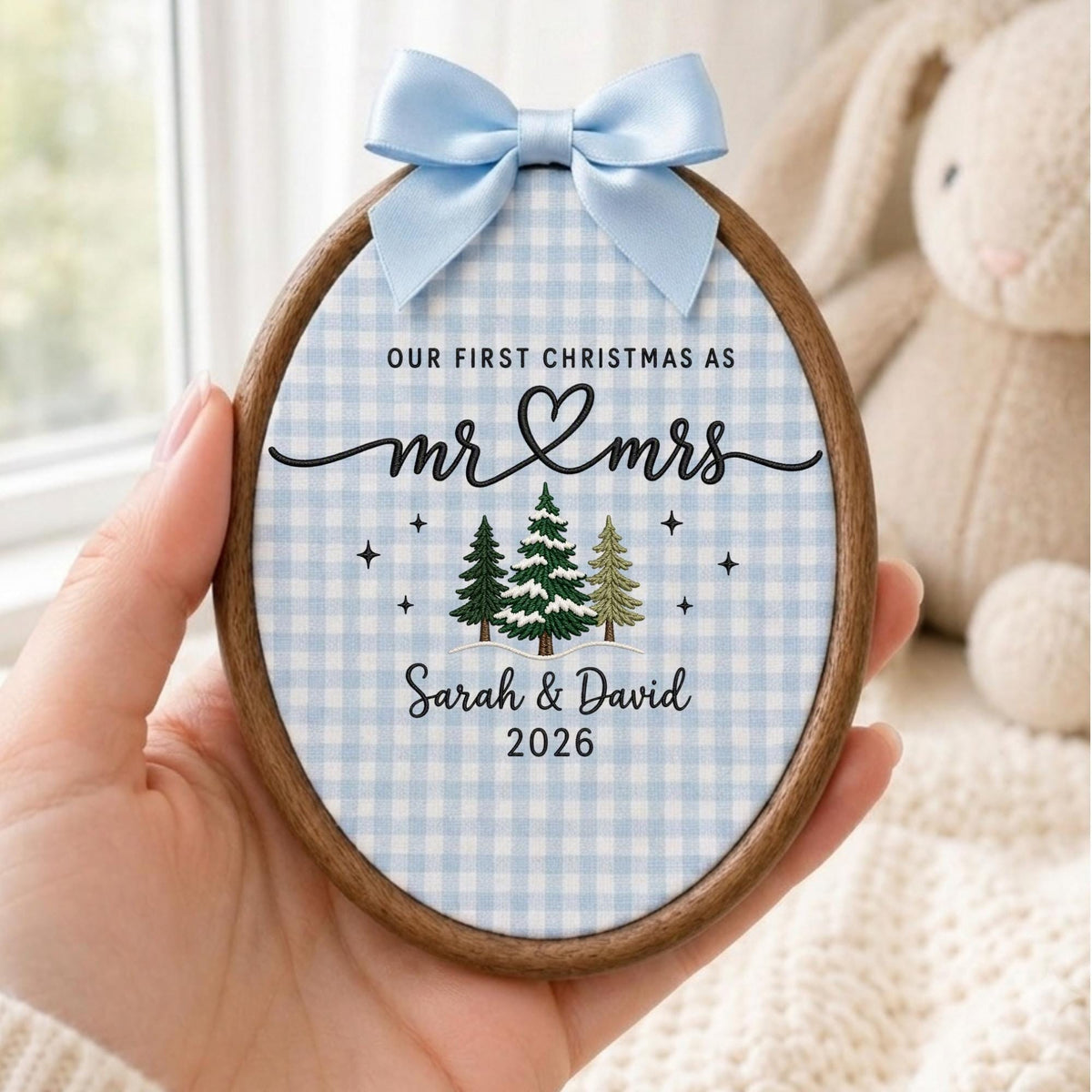 May include: An oval wooden embroidery hoop ornament featuring a light blue and white gingham fabric background. It displays the text "OUR FIRST CHRISTMAS AS mr &amp; mrs", "Sarah &amp; David", and "2026" with embroidered pine trees and a light blue satin bow.