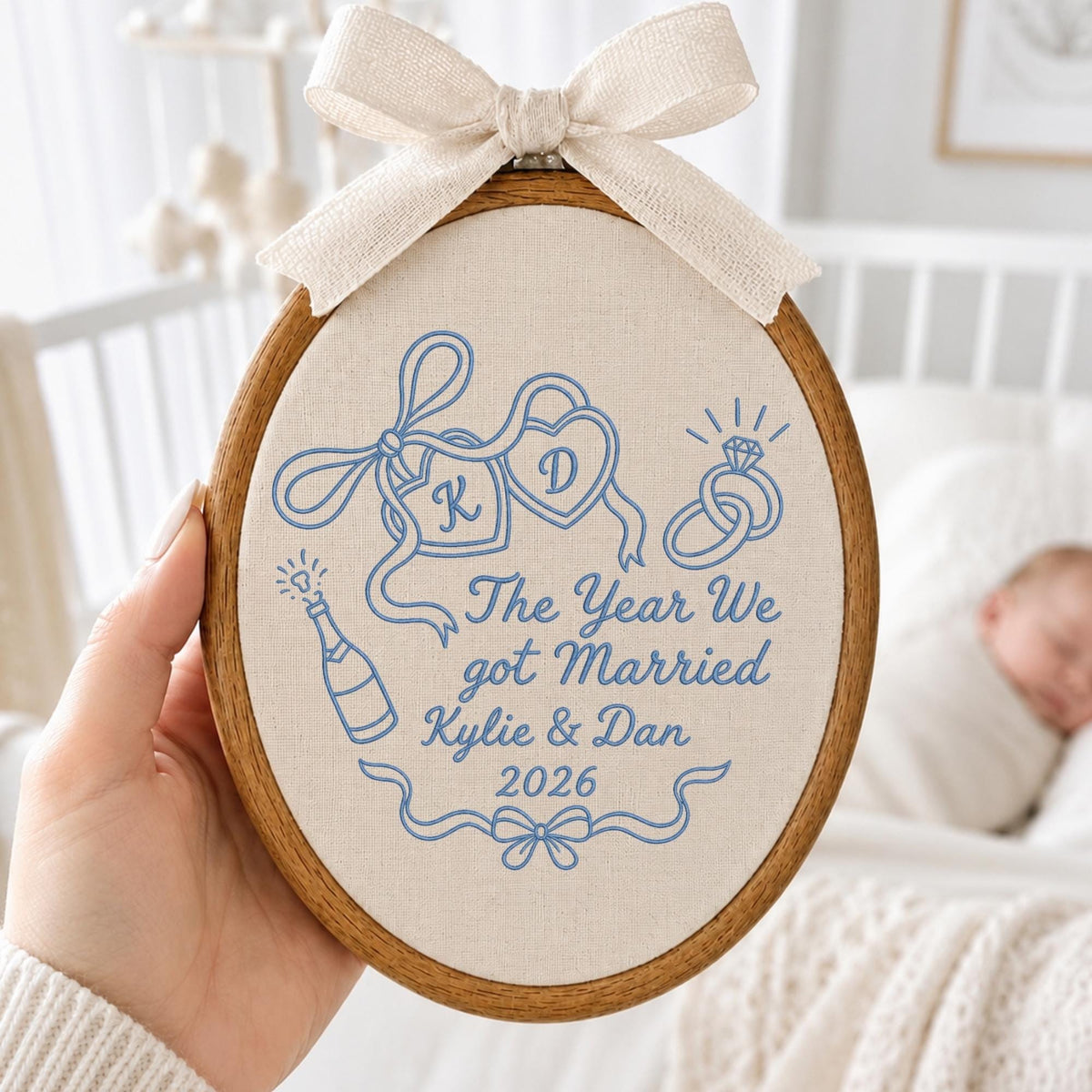 May include: An oval wooden embroidery hoop containing cream-colored fabric with blue stitched designs. The embroidery features two hearts with initials K and D, wedding rings, a champagne bottle, and the text The Year We got Married Kylie &amp; Dan 2026. A beige fabric bow is attached to the top.