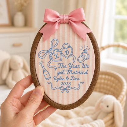 May include: An oval wooden embroidery hoop containing light pink and white striped fabric with a pink satin bow at the top. The fabric features blue embroidered text reading The Year We got Married Kylie &amp; Dan 2026, two hearts with K and D, rings, and a champagne bottle.