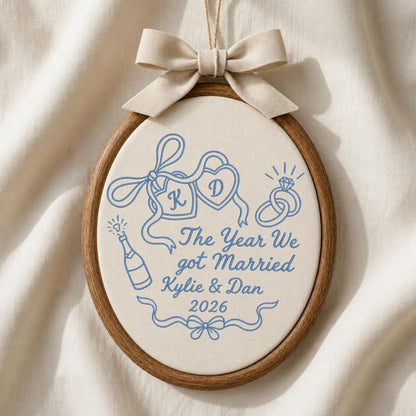 May include: An oval wooden embroidery hoop containing cream fabric with blue stitched designs including two hearts with letters K and D, a ring, a champagne bottle, and the text The Year We got Married Kylie &amp; Dan 2026, topped with a beige fabric bow.