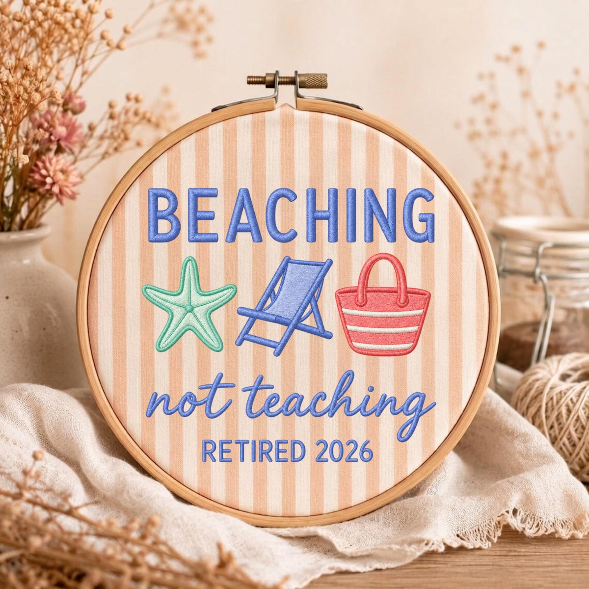 May include: A round wooden embroidery hoop containing fabric with vertical peach and white stripes. The embroidery features the blue text BEACHING not teaching, the year RETIRED 2026, a mint green starfish, a blue beach chair, and a red beach bag.