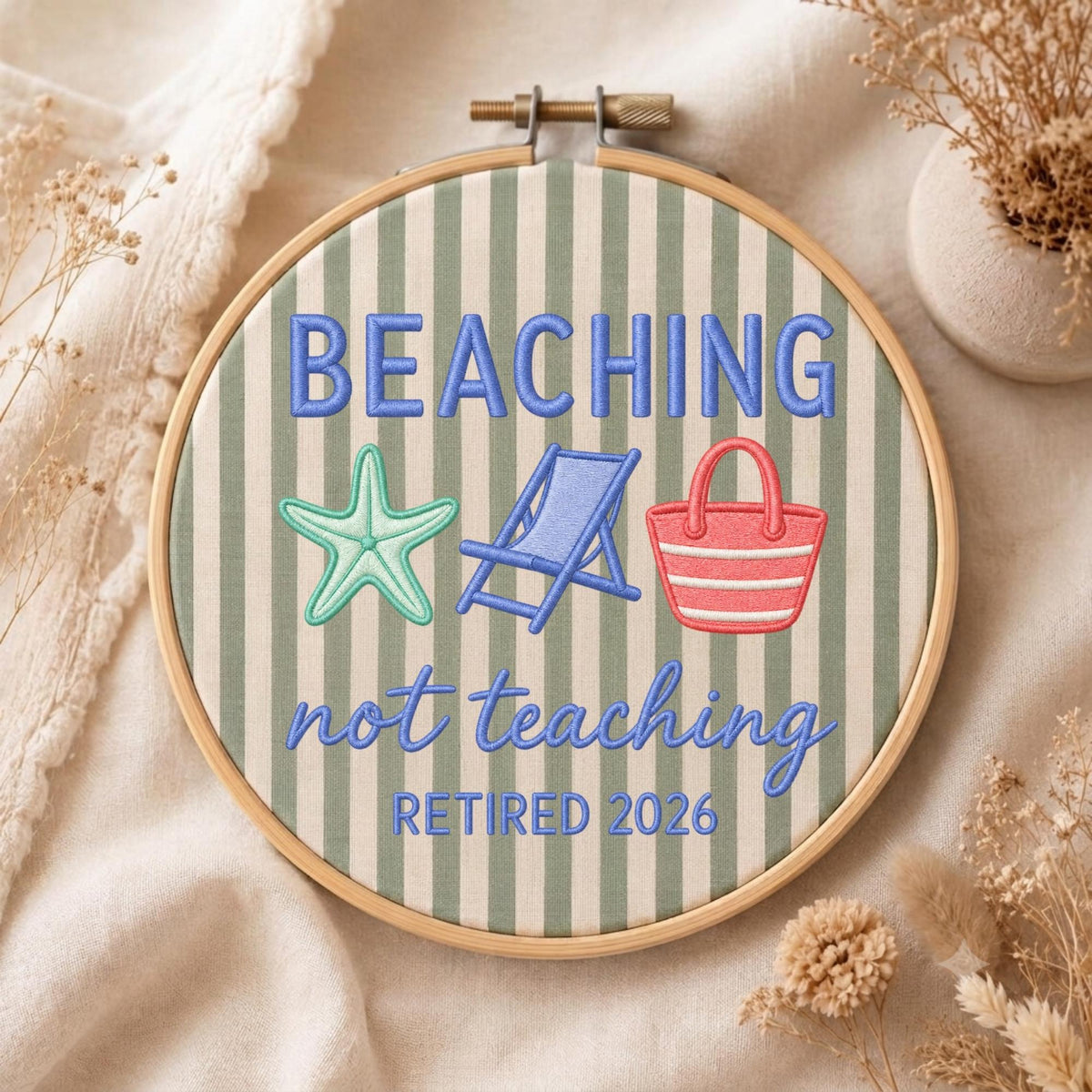 May include: A circular wooden embroidery hoop containing fabric with vertical sage green and white stripes. The embroidery features the blue text BEACHING not teaching RETIRED 2026, a light green starfish, a blue beach chair, and a red beach bag.