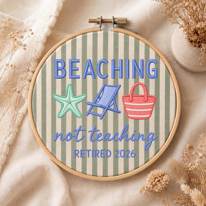 May include: A circular wooden embroidery hoop containing fabric with vertical sage green and white stripes. The embroidery features the blue text BEACHING not teaching RETIRED 2026, a light green starfish, a blue beach chair, and a red beach bag.