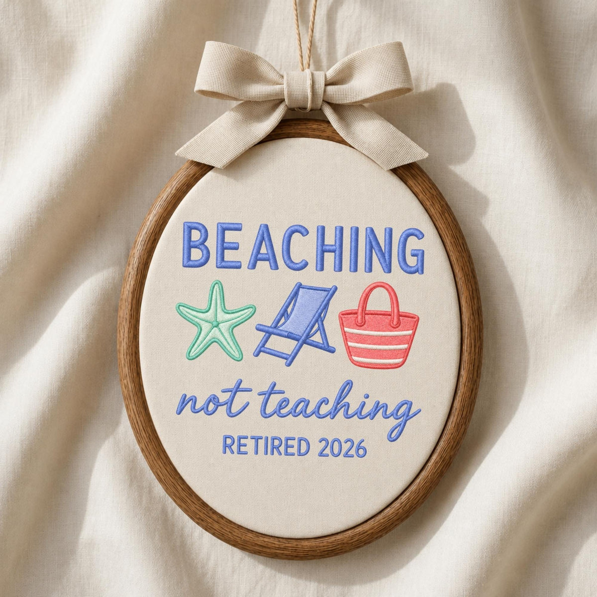May include: An oval embroidery hoop containing off-white fabric with the embroidered text BEACHING not teaching RETIRED 2026. The design includes a light green starfish, a blue beach chair, and a red beach bag. A beige ribbon bow is tied at the top.