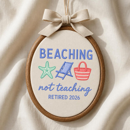 May include: An oval embroidery hoop containing off-white fabric with the embroidered text BEACHING not teaching RETIRED 2026. The design includes a light green starfish, a blue beach chair, and a red beach bag. A beige ribbon bow is tied at the top.