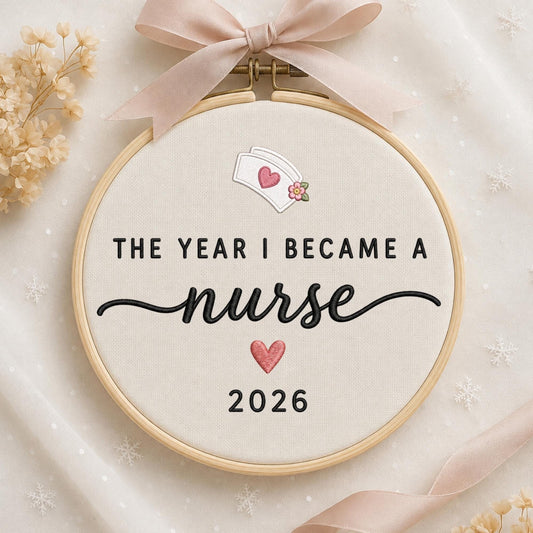 May include: A circular wooden embroidery hoop containing cream-colored fabric with the black embroidered text THE YEAR I BECAME A nurse, a white nurse cap with a pink heart, a small pink heart, and the year 2026, topped with a light pink ribbon.