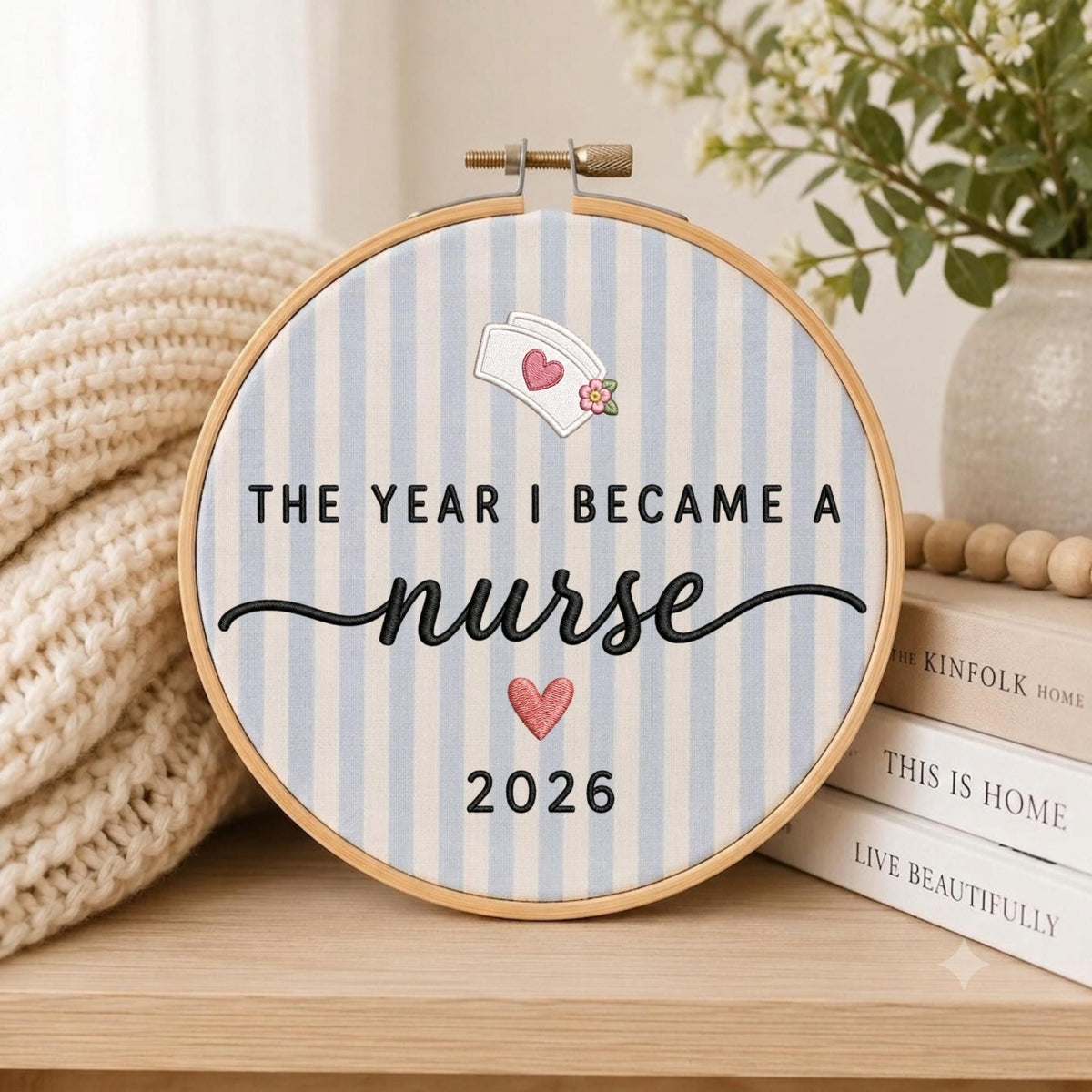 May include: A circular embroidery hoop containing light blue and white striped fabric. The text reads THE YEAR I BECAME A nurse 2026. Above the text is a nurse cap icon with a pink heart, and below the text is a pink heart. The frame is wood.
