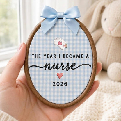 May include: An oval wooden embroidery hoop frame containing fabric with a light blue and white gingham pattern. The text reads THE YEAR I BECAME A nurse 2026, accompanied by an embroidered nurse cap icon and two small pink hearts.