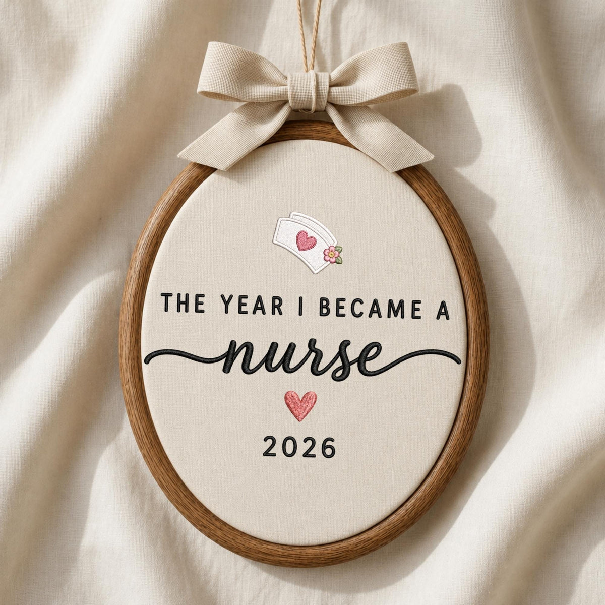 May include: An oval wooden embroidery hoop containing cream-colored fabric with the text THE YEAR I BECAME A nurse 2026. Above the text is a nurse cap icon with a pink heart and flower, and below is a pink heart. A beige ribbon bow is tied at the top.