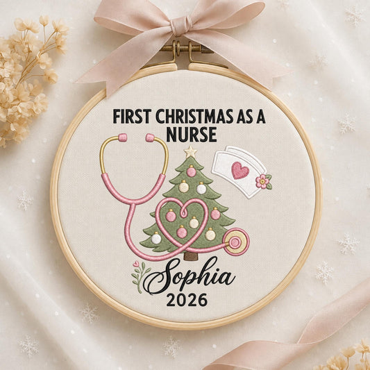 May include: A round wooden embroidery hoop featuring a cream fabric background with the embroidered text FIRST CHRISTMAS AS A NURSE, a green Christmas tree, a pink stethoscope, a nurse cap, and the name Sophia 2026, topped with a light pink ribbon.