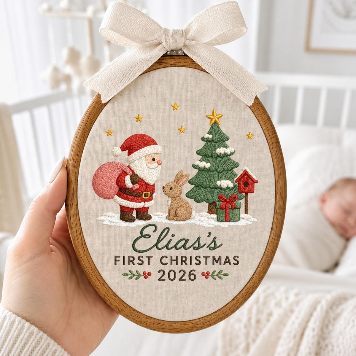 May include: An oval embroidery hoop featuring a cream fabric background with a stitched scene of Santa Claus, a brown rabbit, a green Christmas tree, and a red birdhouse. The text reads Elia's FIRST CHRISTMAS 2026. A beige ribbon bow is at the top.