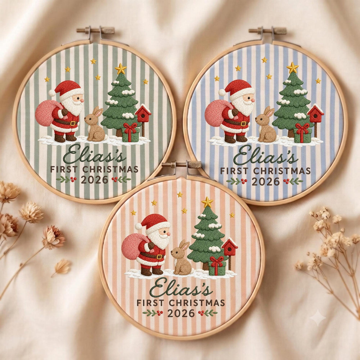 May include: Three circular embroidery hoops featuring a striped background in sage green, light blue, and peach. Each displays a Santa Claus, a rabbit, a Christmas tree, and a birdhouse with the text Elias's FIRST CHRISTMAS 2026.